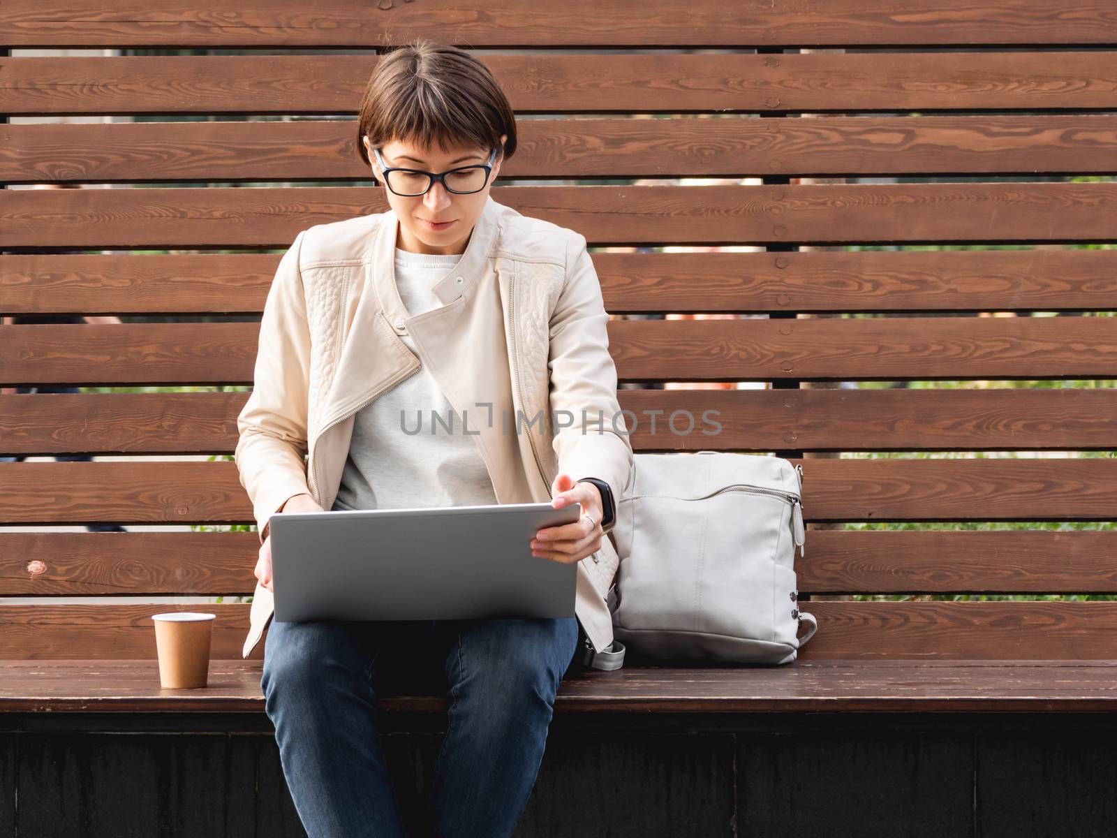 Business woman with short haircut and eyeglasses sits on wooden bench in park with laptop and cardboard cup of coffee. Student learns remotely from outdoors. Modern Internet technologies. Video call.