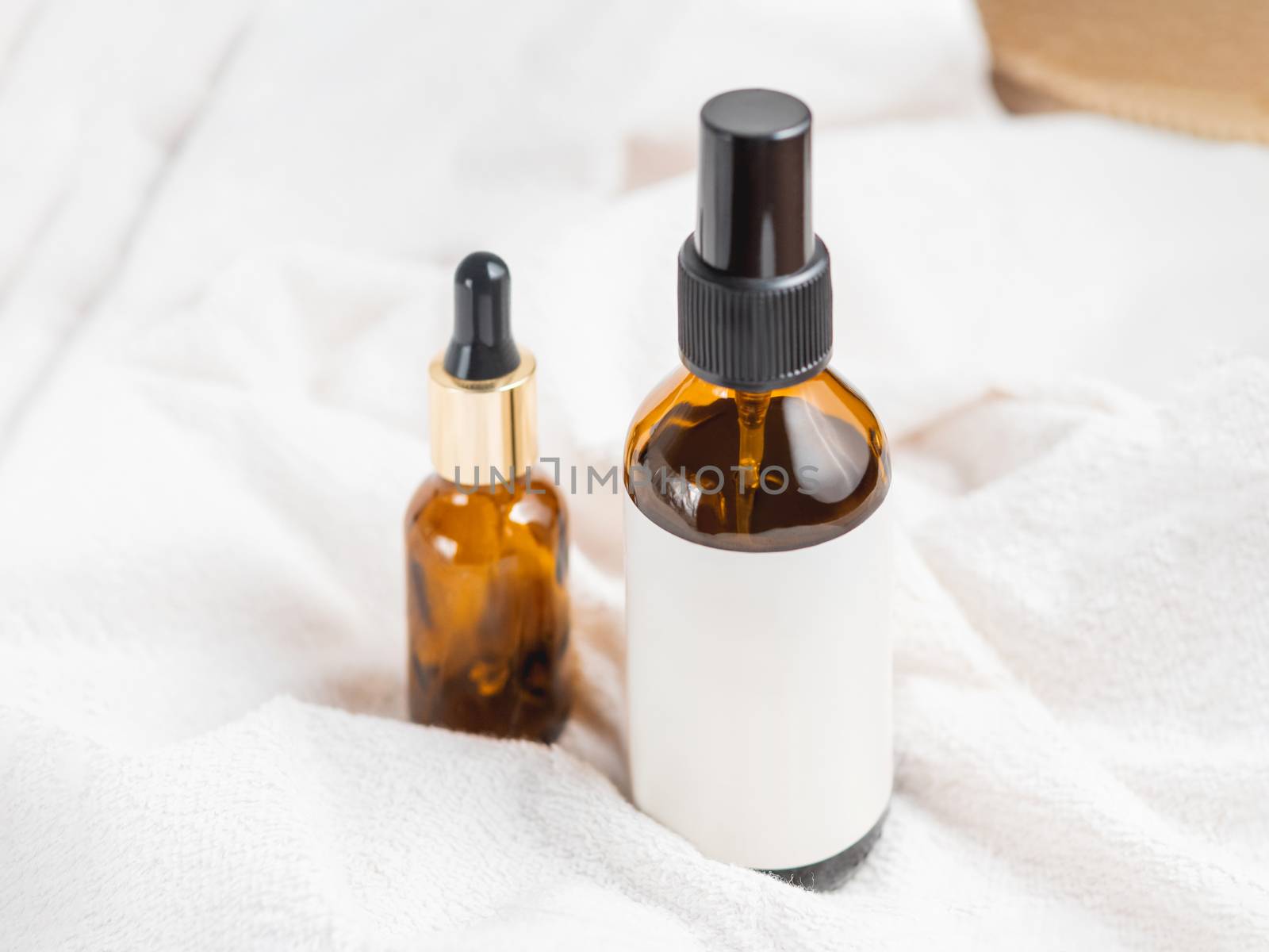 Two glass bottles on white fluffy towel. Essential oil and cosmetic serum with clear labels on crumpled bathrobe. Morning routine.