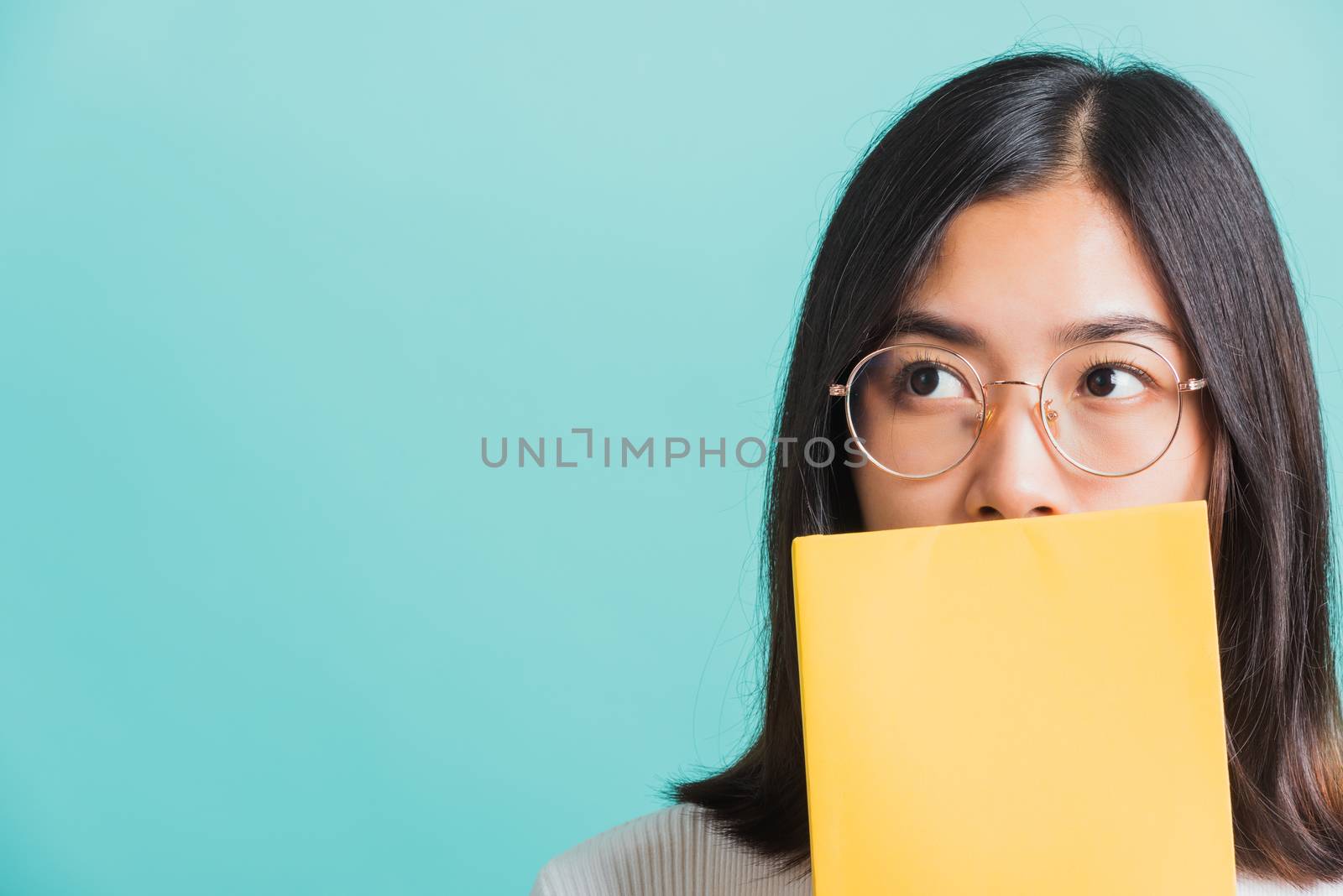 Young beautiful Asian woman hiding behind an open book, Portrait female in glasses is holding and reading a book, studio shot isolated on a blue background, Education concept