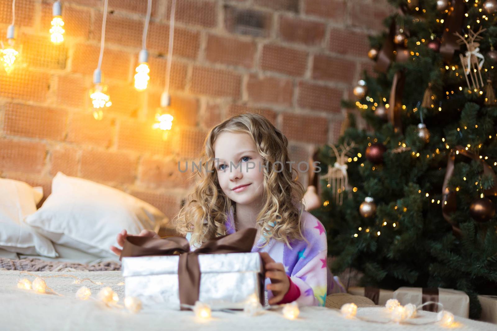 Cute Girl in pajamas lies on bed with a gift box and new year tree and lights behind