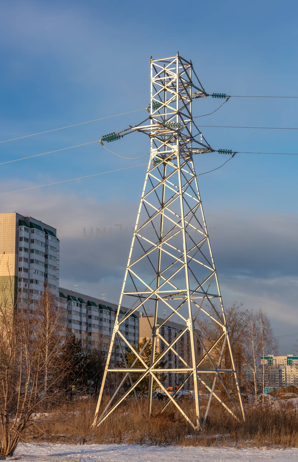 Low angle shot of high voltage electric tower and power lines in the city at sunset. Apartment buildings and blue cloudy sky as a background. Barnaul, Siberia, Russia.