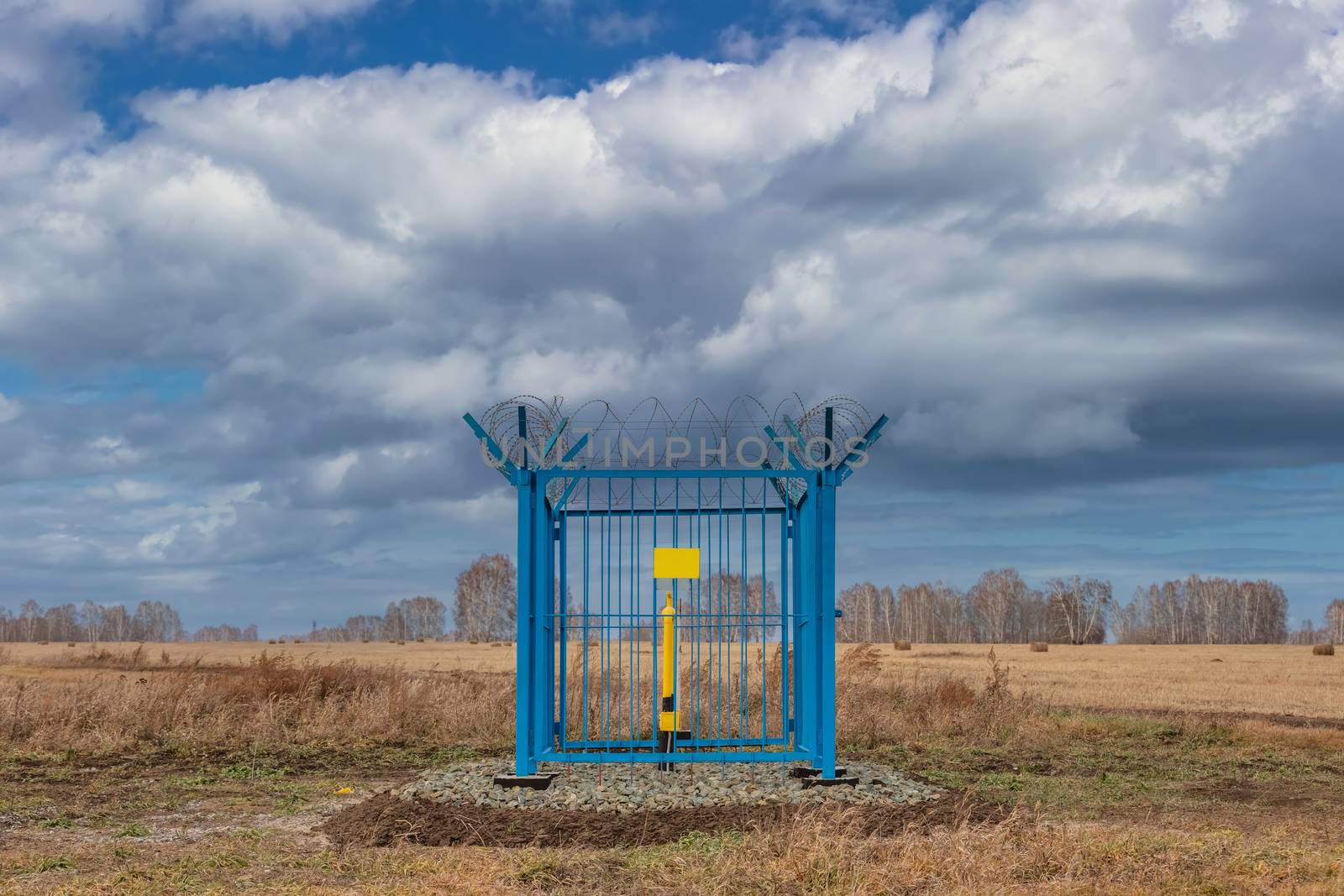 Shot of a caged industrial gas station with barbwire on top of the fence. Yellow gas pipe in the middle of the cage, empty yellow plate on the fence. Blue cloudy sky as a background. Siberia, Russia by DamantisZ