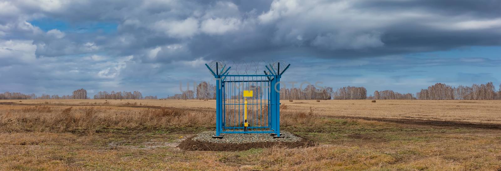 Panoramic shot of a caged industrial gas station with barbwire on top of the fence. Yellow gas pipe in the middle of the cage, empty plate on the fence. Cloudy sky as a background. Siberia, Russia by DamantisZ