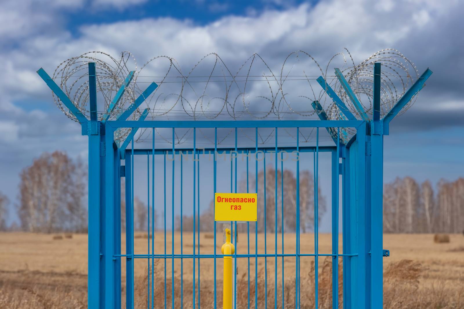 Shot of a caged industrial gas station with barbwire on top of the fence. Yellow gas pipe in the middle of the cage. Yellow plate on the fence reads in Russian - Flammable. Gas. Siberia, Russia.