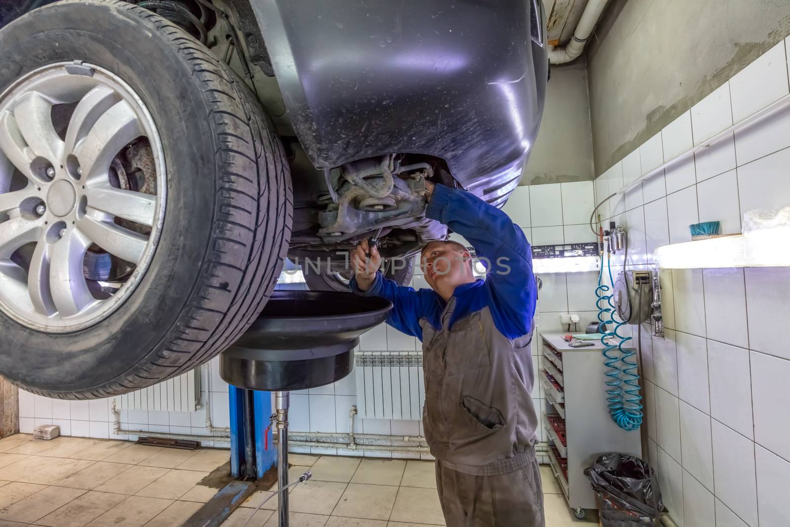 Car mechanic changes oil in a workshop. Mechanic standing under the car and draining oil into a special cannister via funnel by DamantisZ