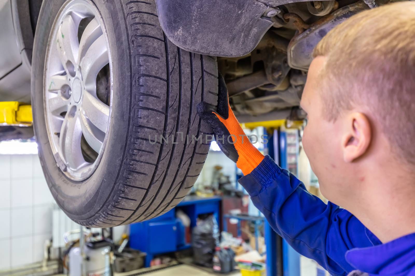 Close-up shot of mechanic checking very attentively surface of a car tire. Mechanic wearing blue coverall and orange gloves. Car is on a hydraulic lift.