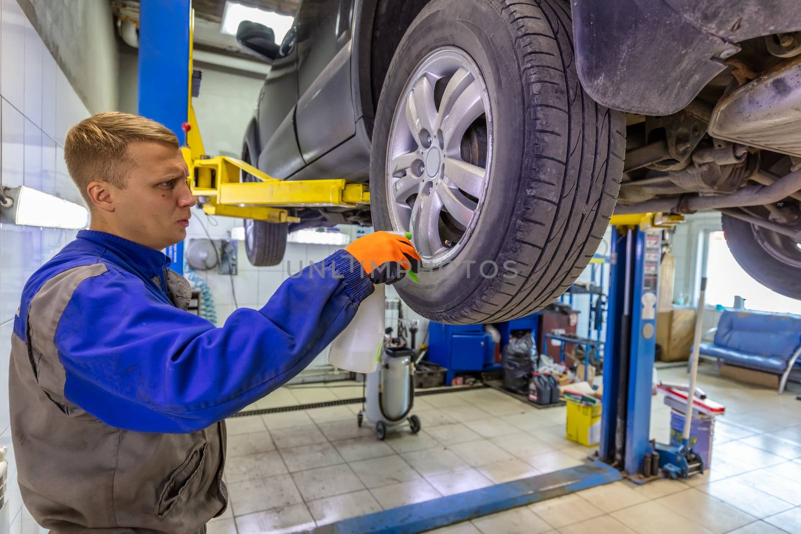 Close-up shot of mechanic checking very attentively surface of a car tire using a spray bottle. Mechanic wearing blue coverall and orange gloves. Car is on a hydraulic lift.