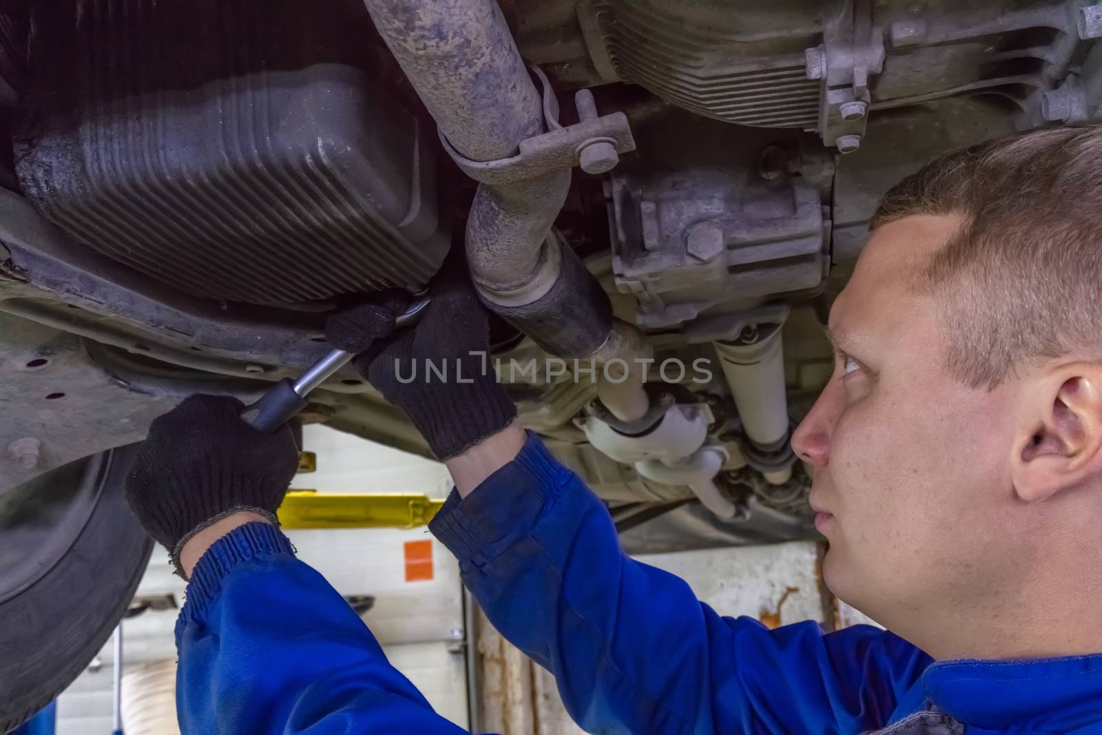 A close-up shot of a mechanic working under a car at the garage. Technician wearing blue coverall and using a wrench. Car is on a hydraulic lift.