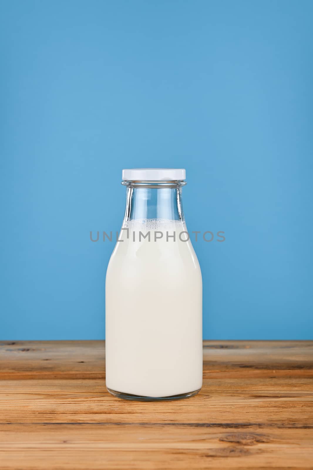 Close up one glass bottle full of fresh milk on wooden table over blue background, low angle, side view