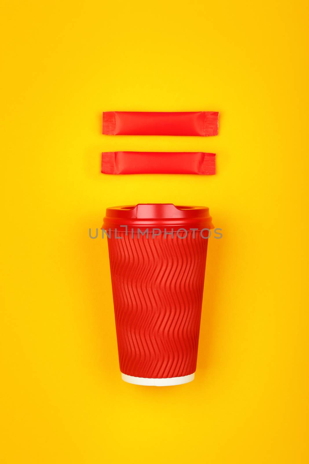 Close up one crimped disposable red paper takeaway cup and two sugar stick sachet over vivid yellow background, flat lay, elevated top view, directly above