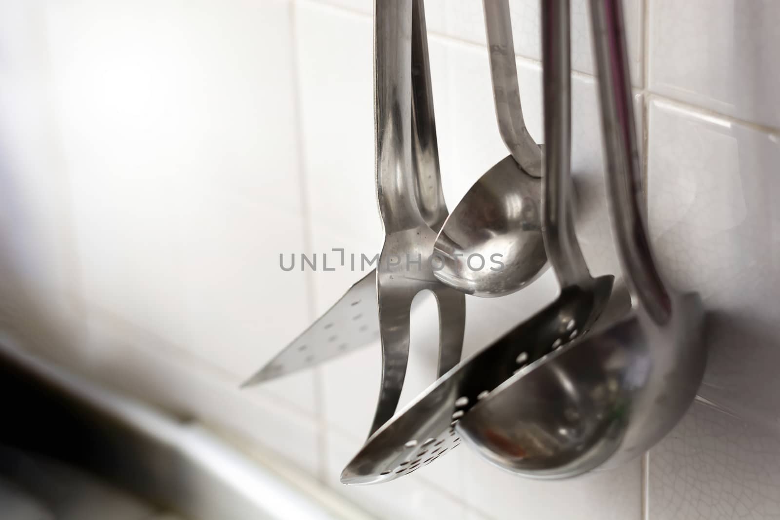 A skimmer, two forks and two metal ladles hanging on the tiled kitchen wall. Metal kitchen utensils