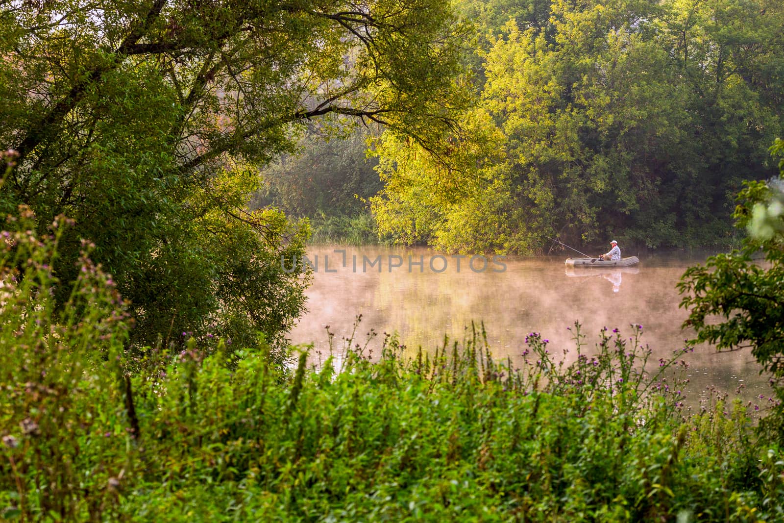 TULA, RUSSIA - AUGUST 12, 2013: A man fishing on river at inflatable boat at foggy summer morning under green foliage. by z1b