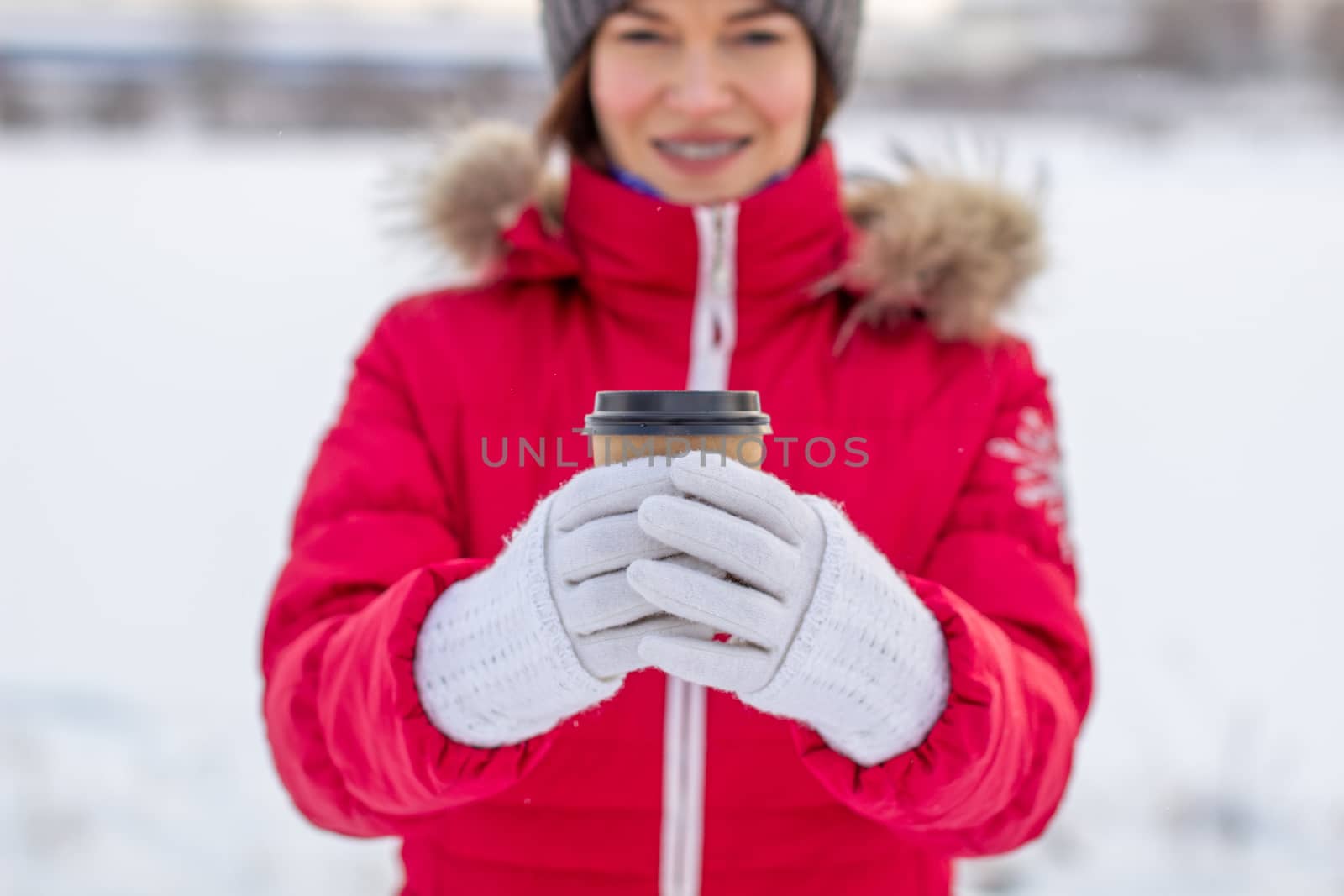 A young woman in a red jacket in winter holds a glass of hot coffee or tea. A snowy winter and a hot drink to keep you warm.