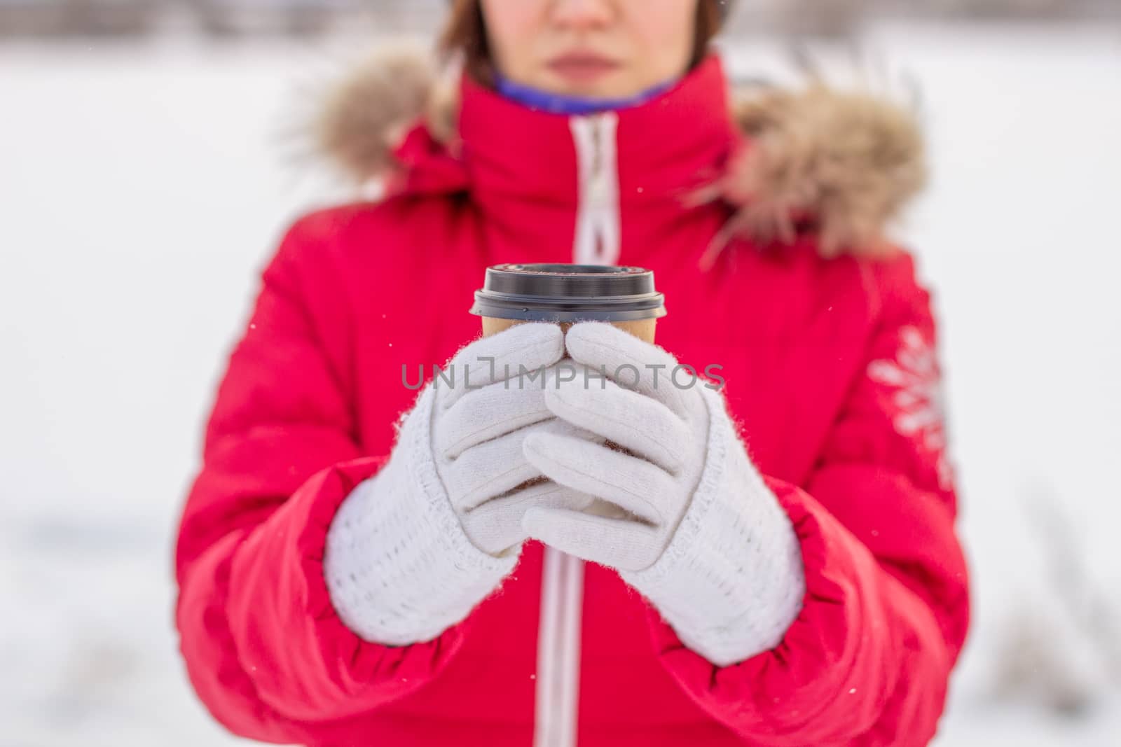 A young woman in a red jacket in winter holds a glass of hot coffee or tea. A snowy winter and a hot drink to keep you warm. A glass of coffee in winter.