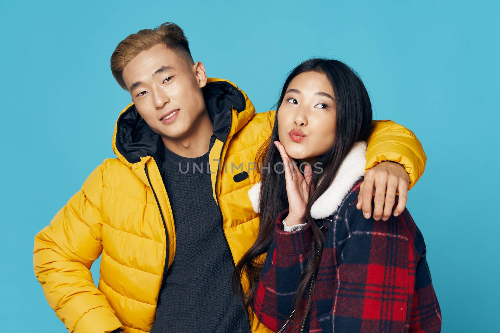 Fashionable young couple of Asian appearance in winter clothes hugs studio blue background