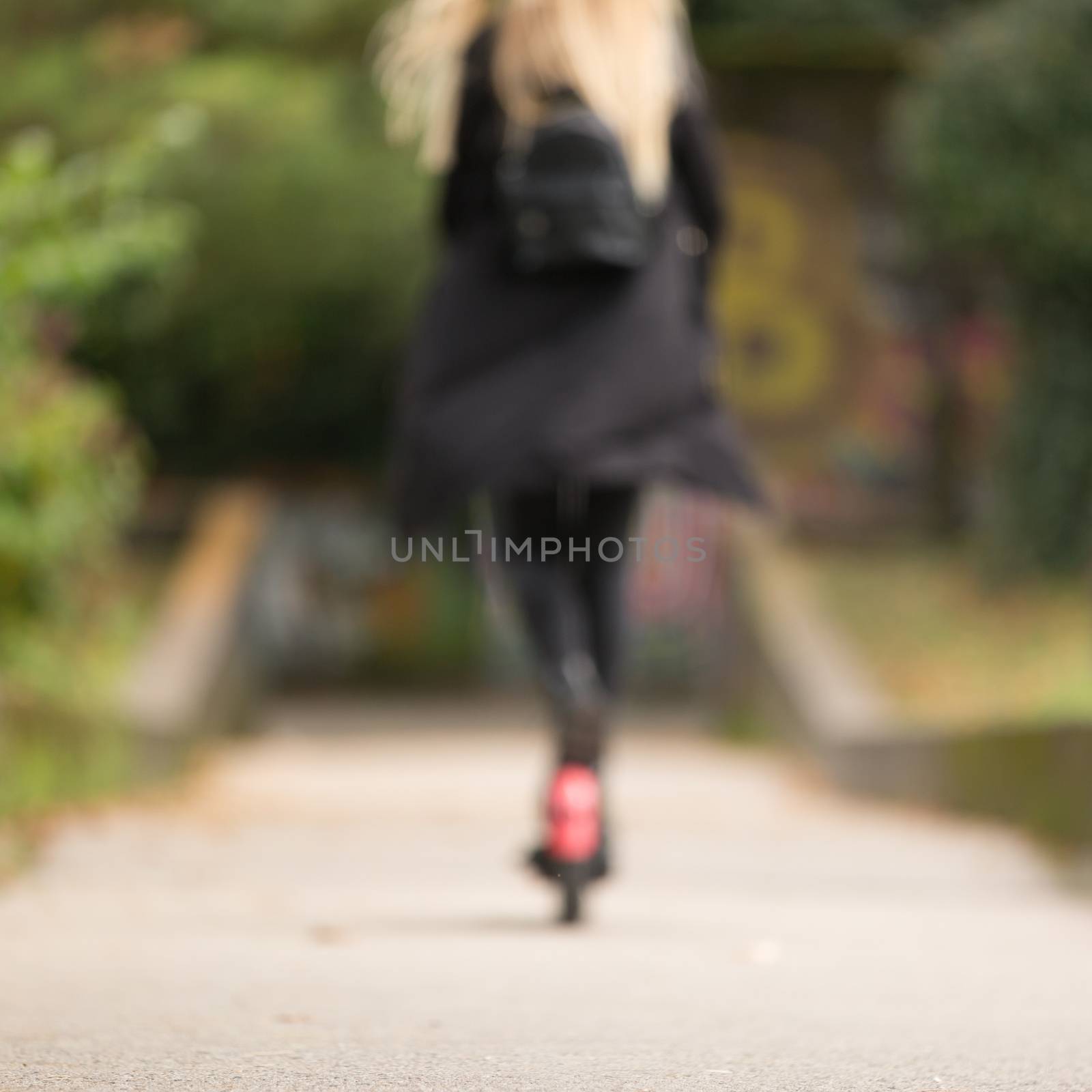 Defocused rear view of casual caucasian teenager riding urban electric scooter in city park during covid pandemic. Urban mobility concept.