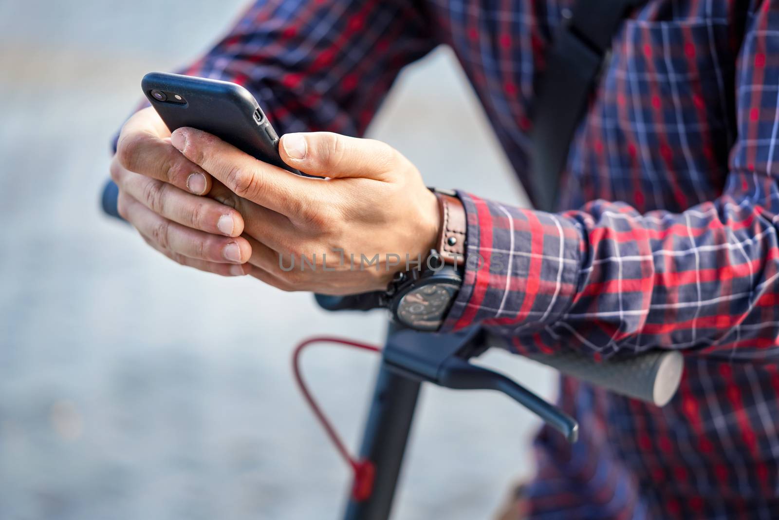 Young man wearing shirt leaning on electric scooter handlebar, holding mobile smartphone in his hands, closeup detail by Ivanko