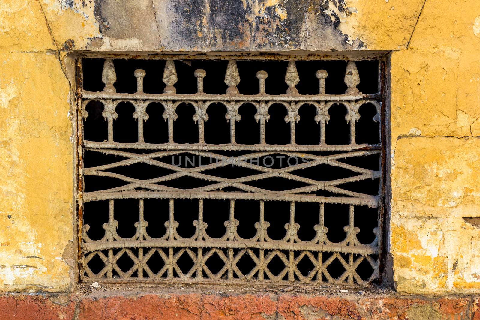 Old yellow wall with closed window with old painted iron grid front
