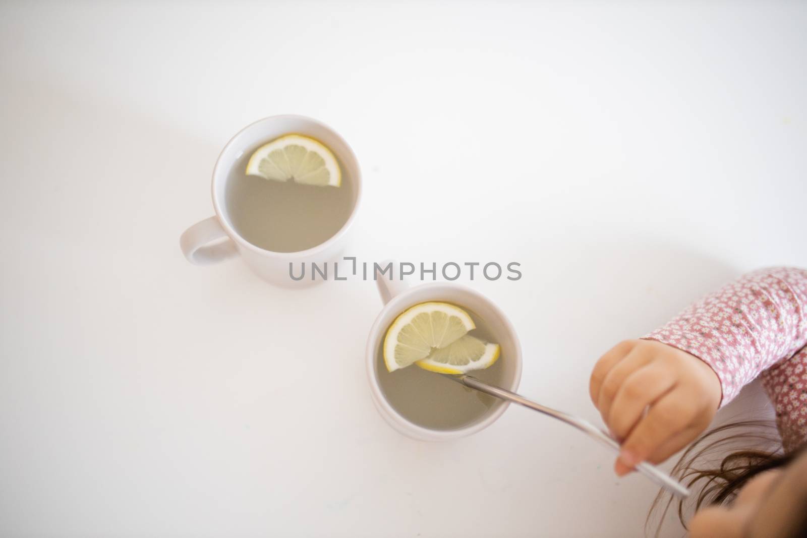 Little girl at table from above drinking cup of lemon tea through a straw. Young child sipping lemon-flavored drink. Sweet citric beverages