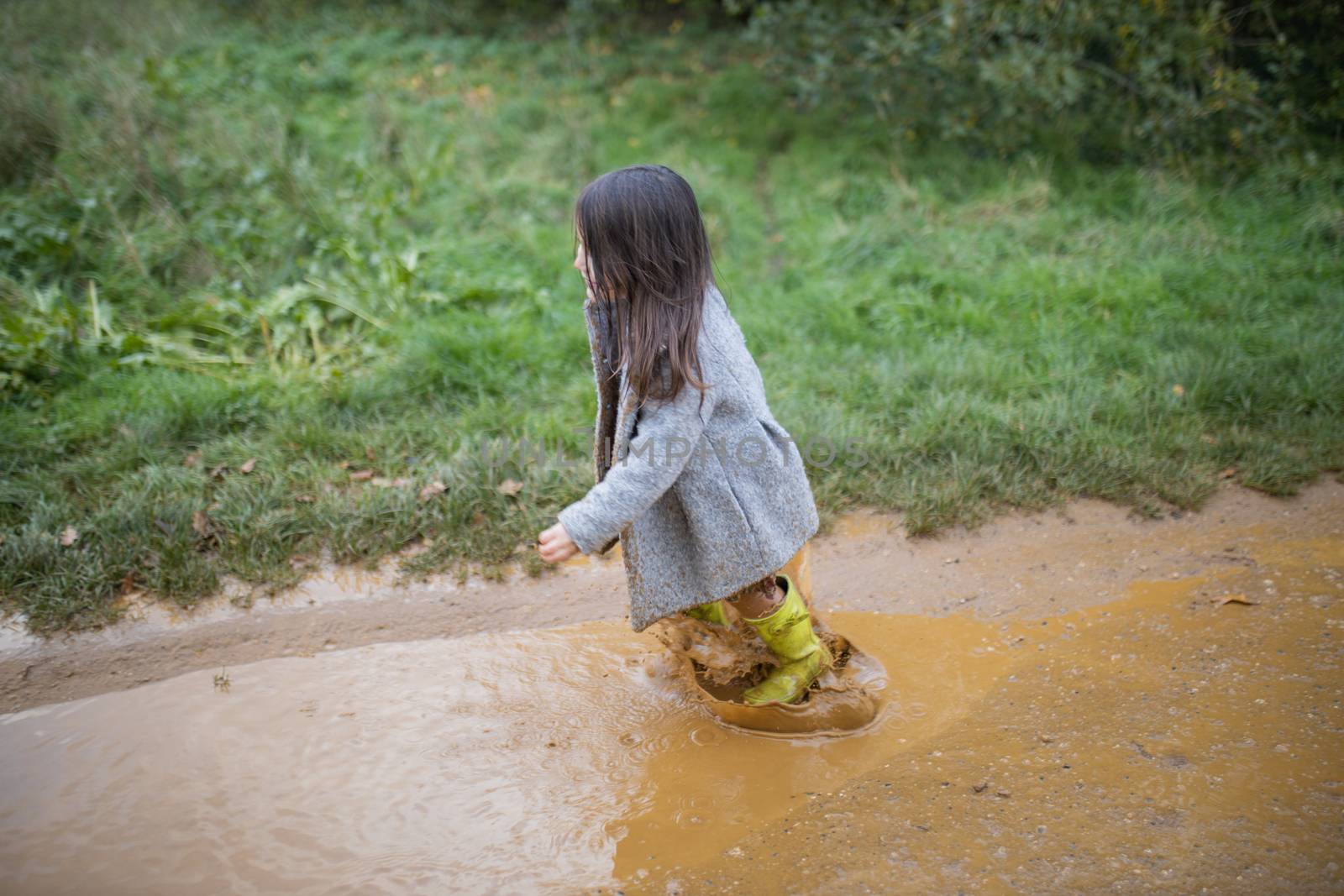 Happy little girl on dirt road joyfully jumping and splashing in muddy puddle. Young child having fun and getting dirty in puddles. Kids playing outside