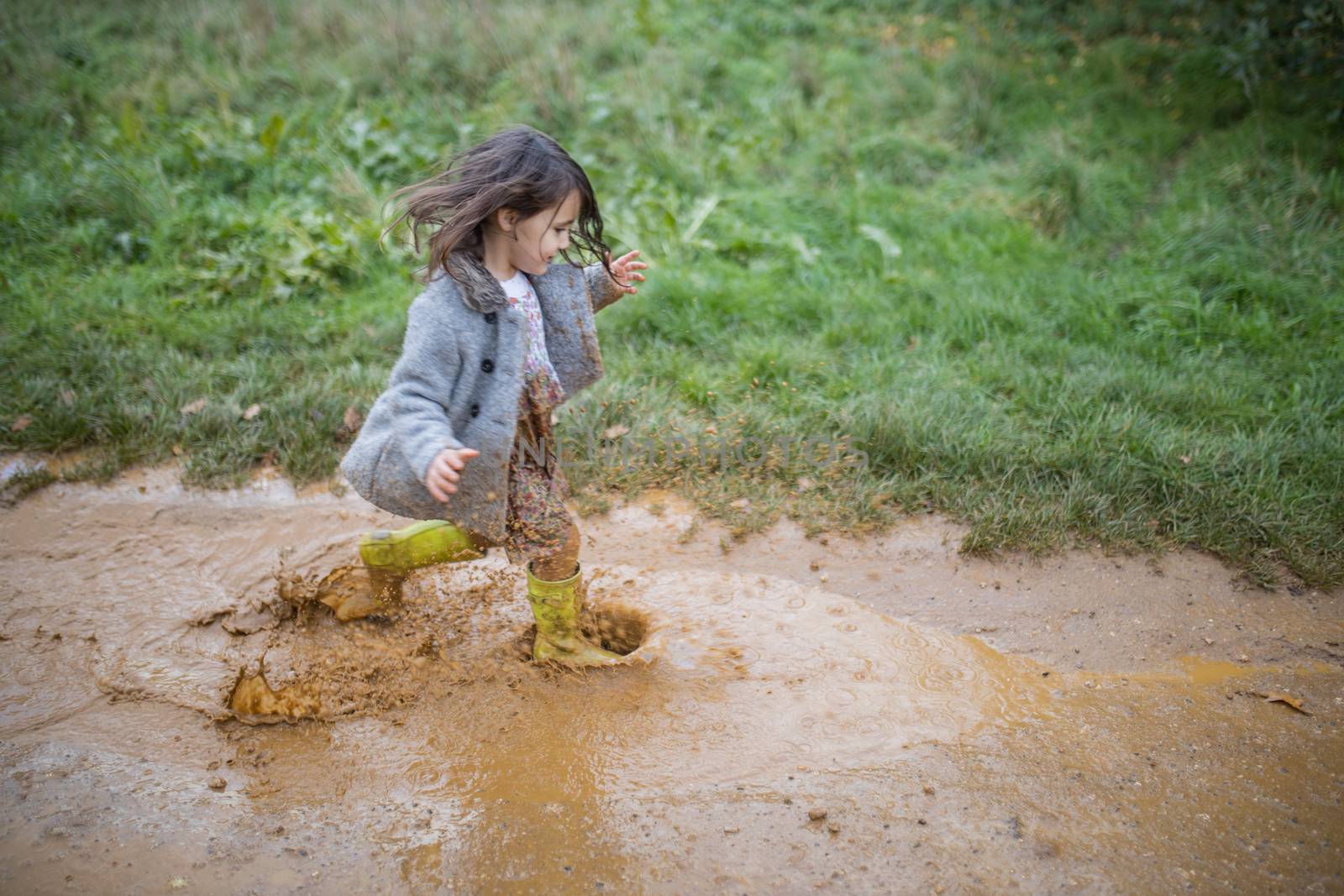 Happy little girl joyfully splashing and running through a muddy puddle. Young child having fun and getting dirty in puddles. Kids playing outside