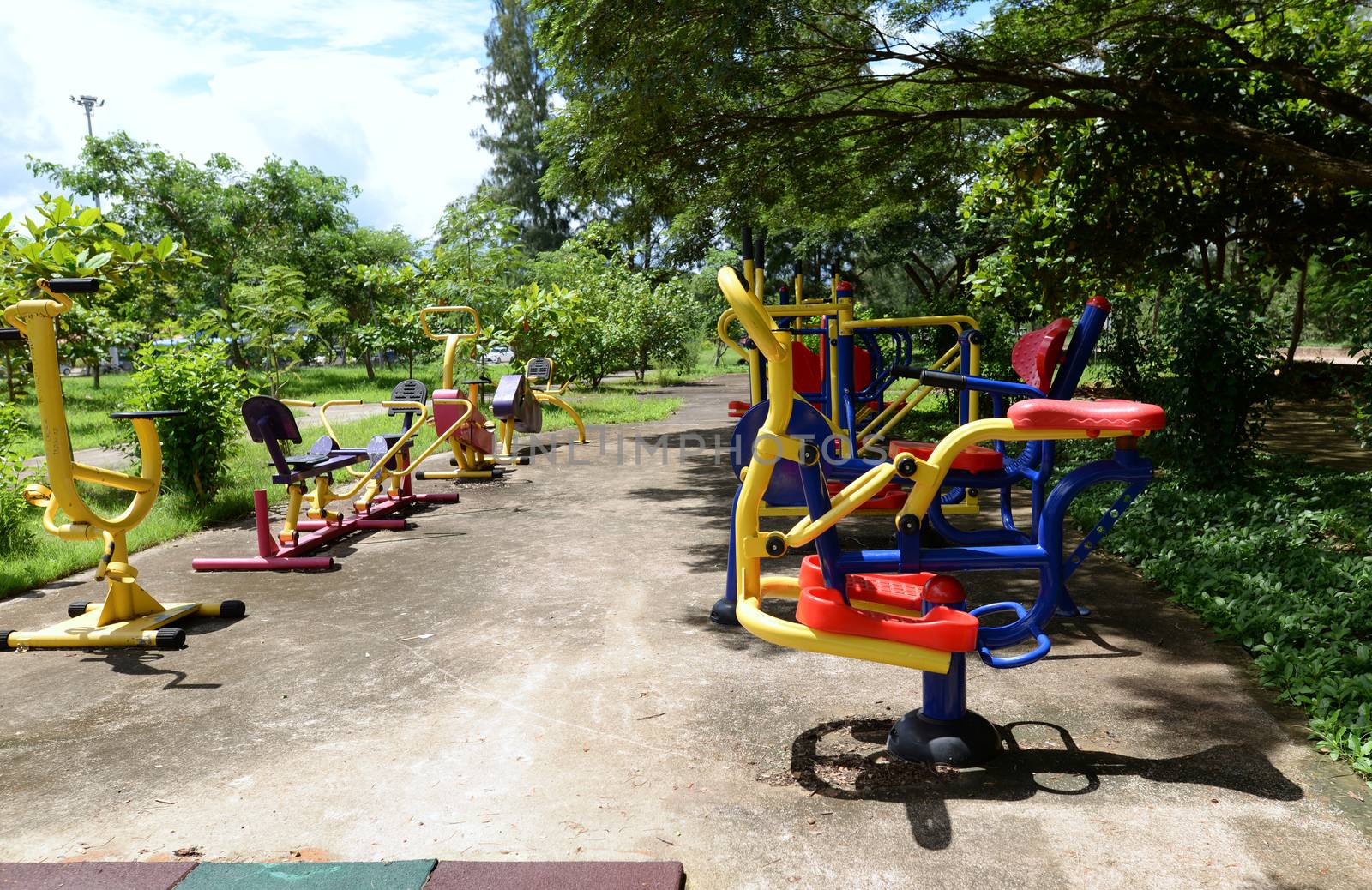 The colorful outdoor exercise machines in the park are lush many green trees.