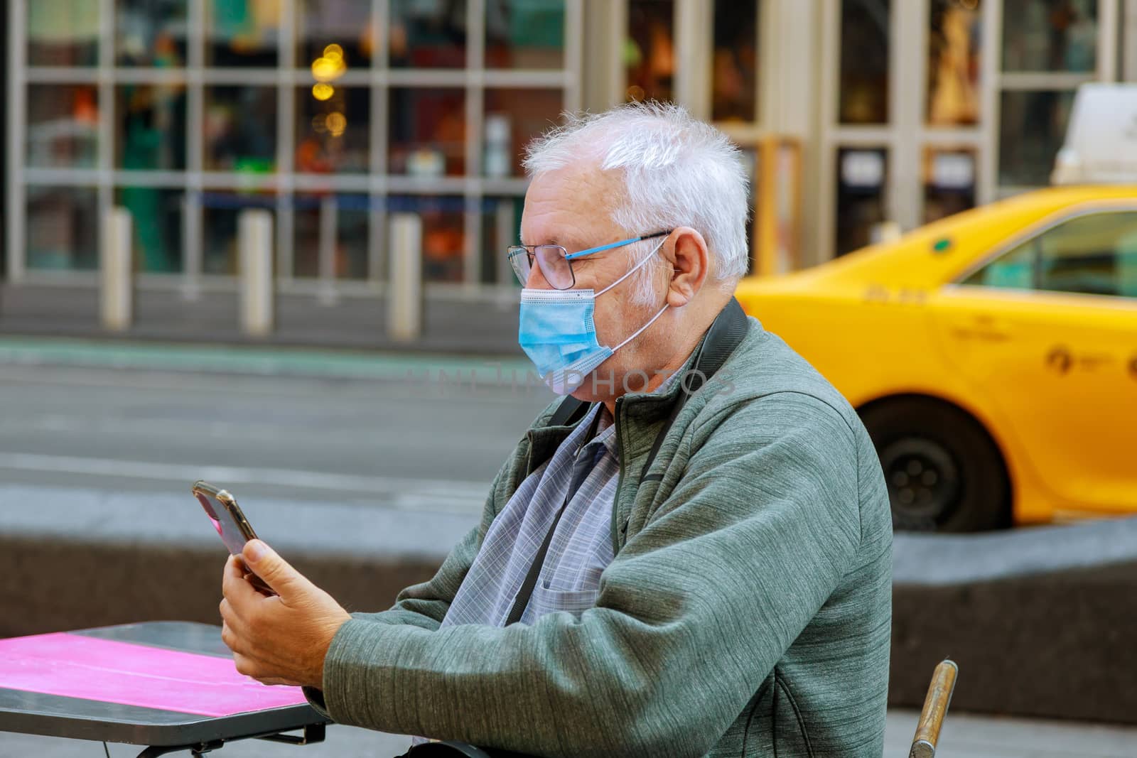 Old man using his mobile phone while walking down the street mask to protect from the coronavirus during the pandemic traveling New York USA