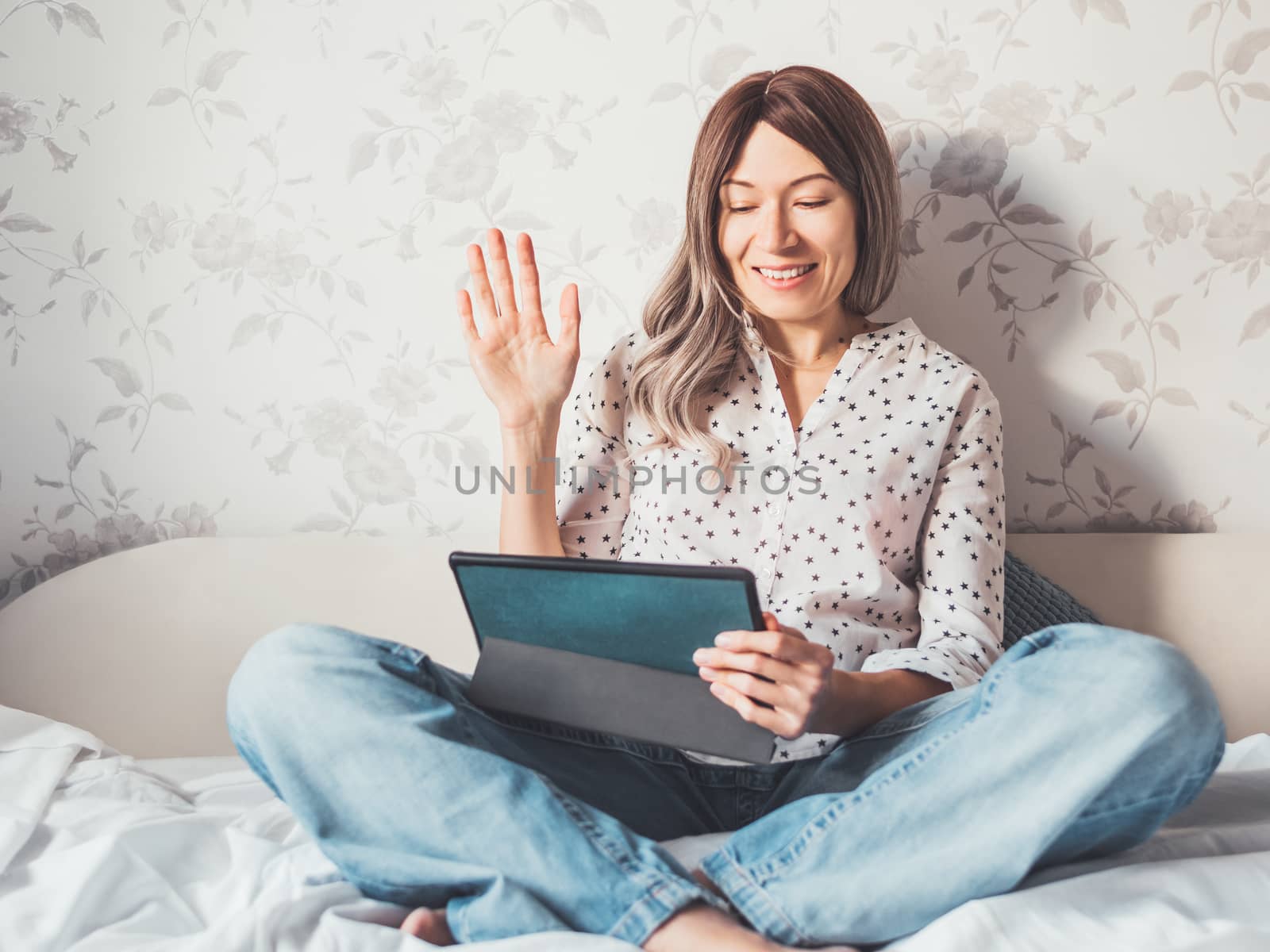 Woman sits on bed with tablet PC. She greets somebody via online video call or conference. Distant learning, remote education. Self isolation during quarantine.