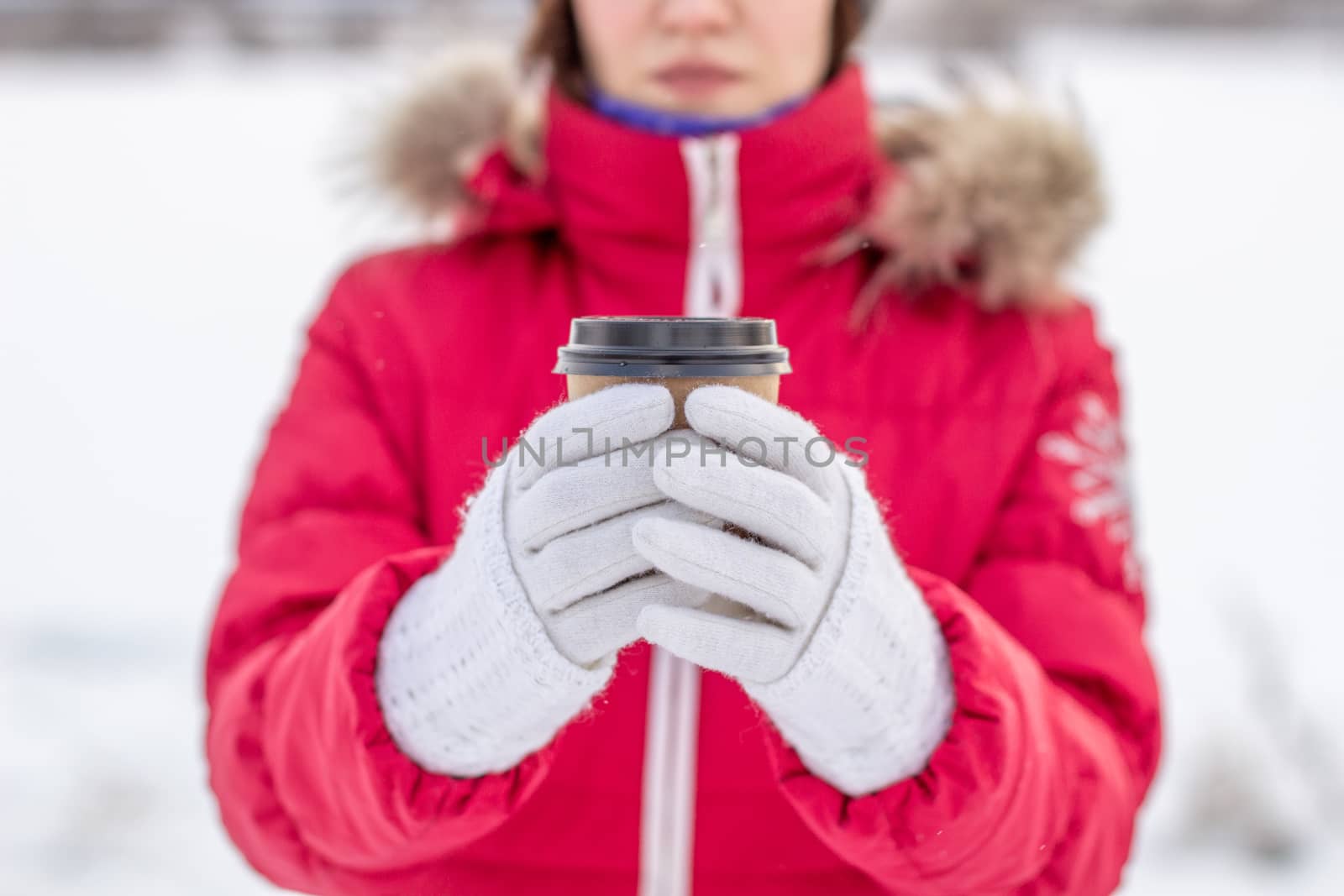 A young woman in a red jacket in winter holds a glass of hot coffee or tea. A snowy winter and a hot drink to keep you warm. A glass of coffee in winter.