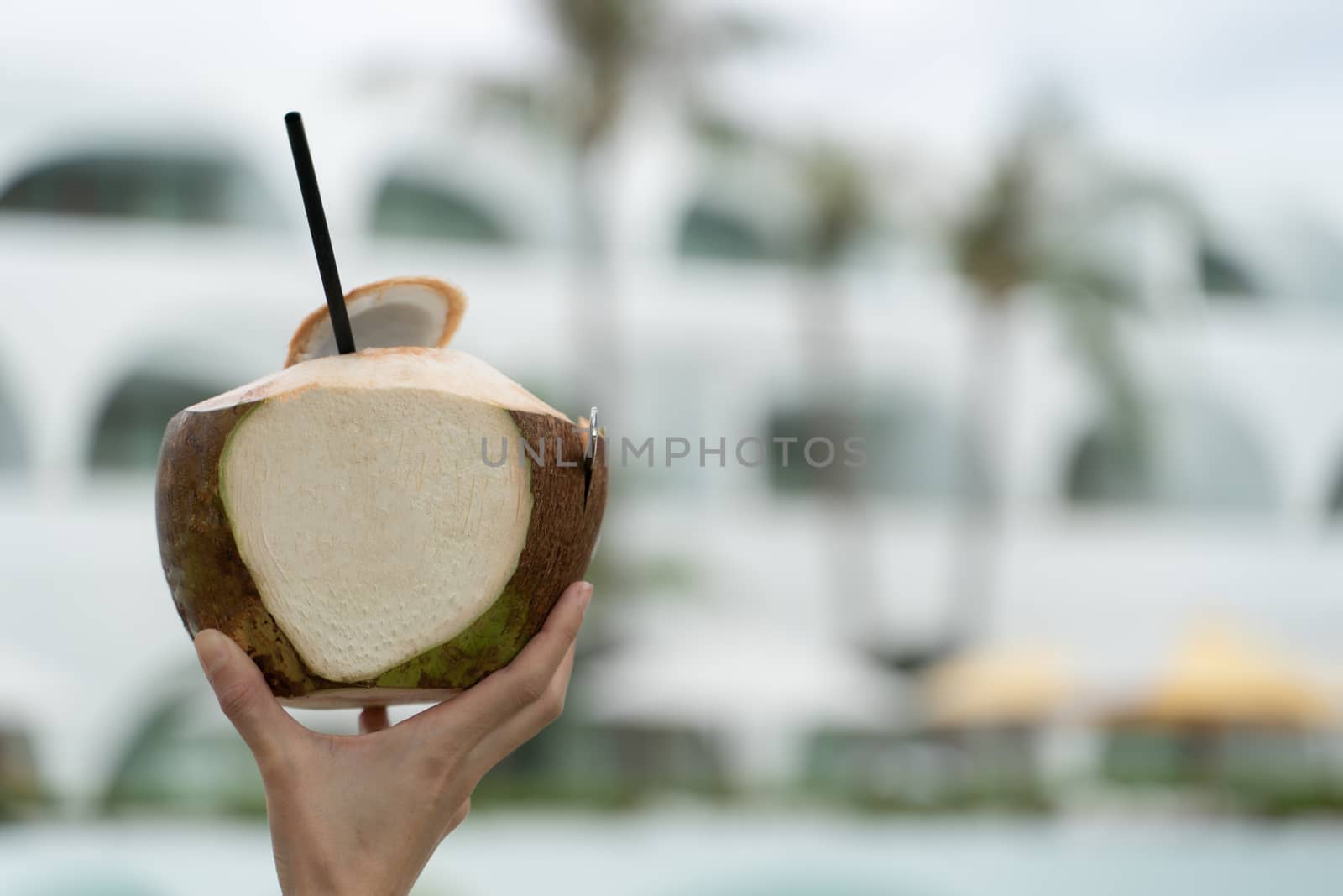 Fresh young coconut juice with straw, hand holding over blurred background.