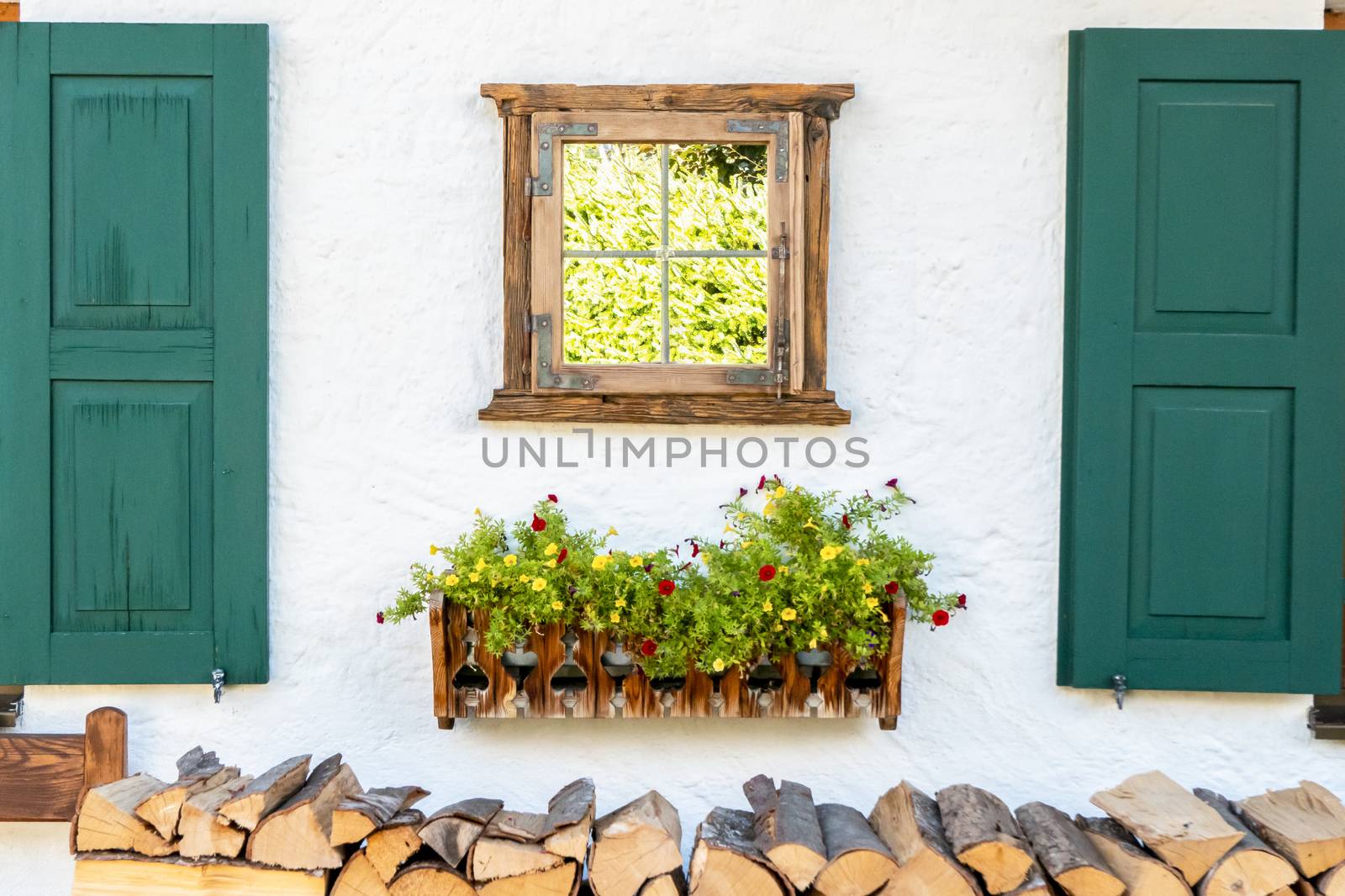 A house wall with a window and shutters, under it a flower box and stacked firewood, which is arranged visually beautiful and decorative.