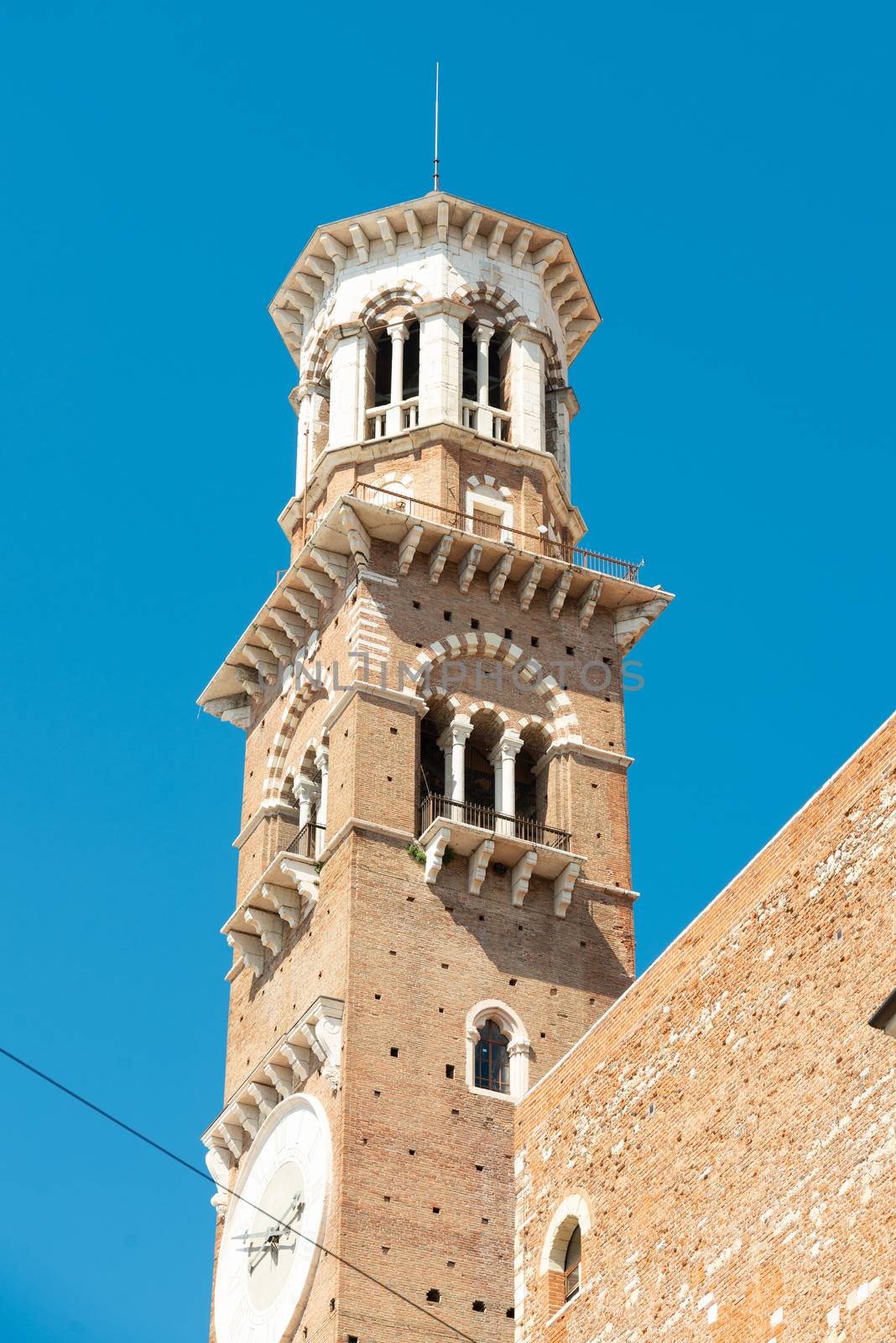 A church tower in Verona photographed from the outside and diagonally below made of stone, a clock, with several ledges in front of a blue sky
