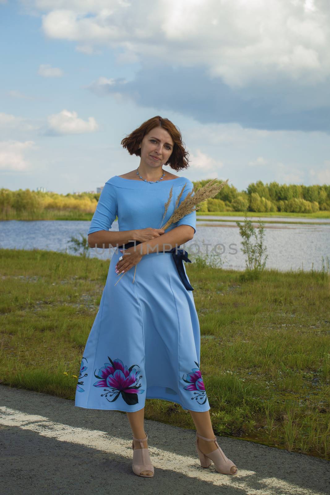 Woman in blue dress with bouquet of flowers. Stands on road against green bank of river. by Essffes