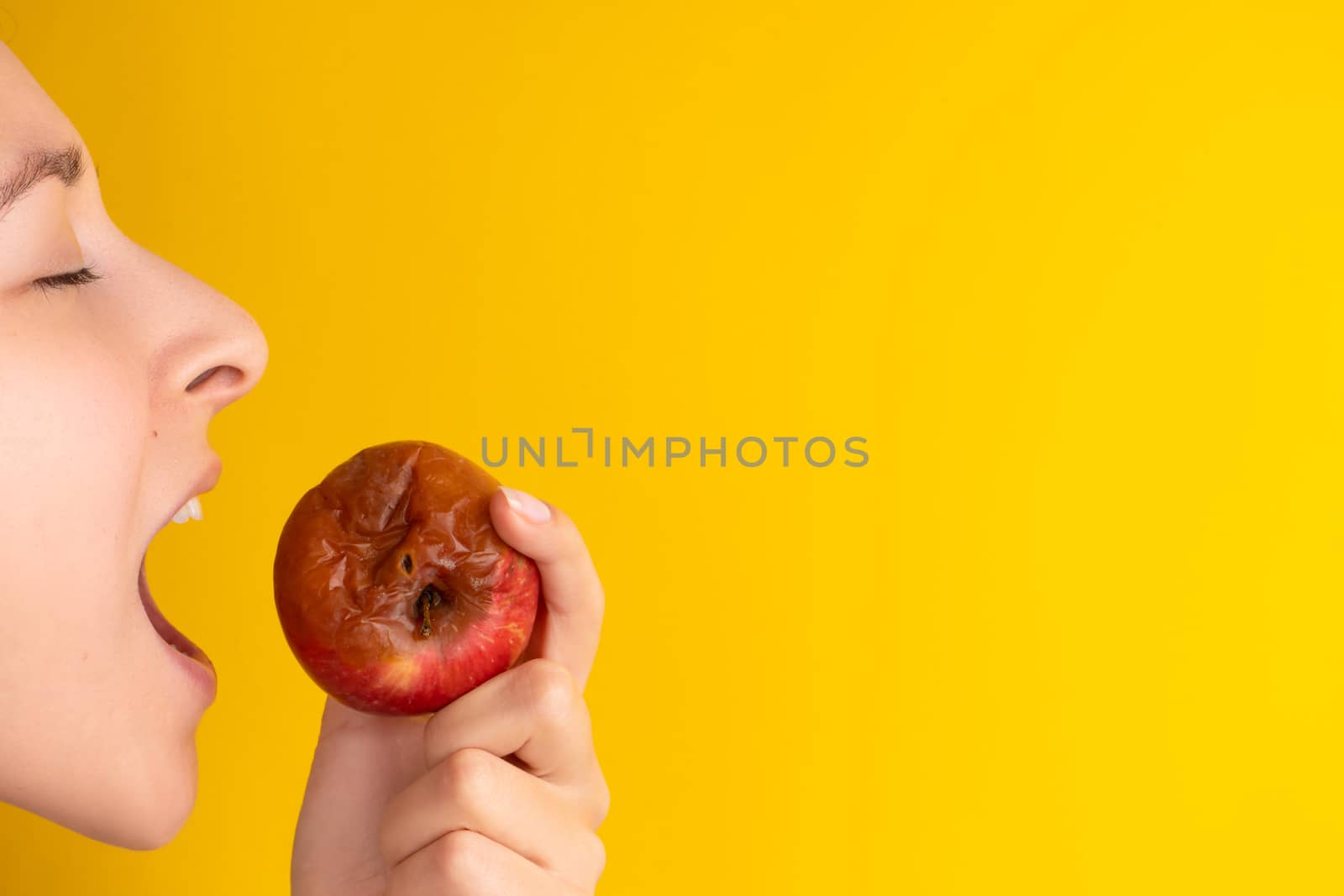 The girl bites a rotten apple with a worm on a yellow background. Expired products, junk food.