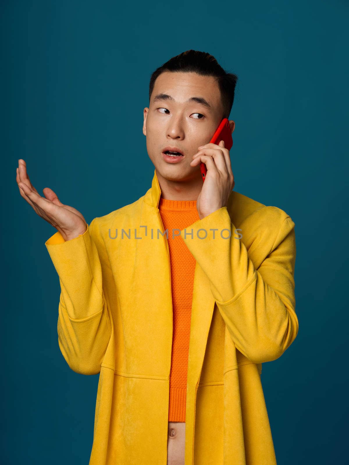 A fashionable man in a yellow jacket and an orange T-shirt is talking on the phone on a blue background by SHOTPRIME