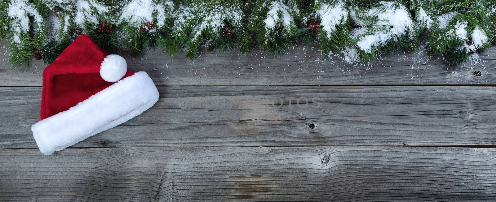 Merry Christmas concept on rustic natural wooden background with snow covered Fir branches plus Santa Claus cap   