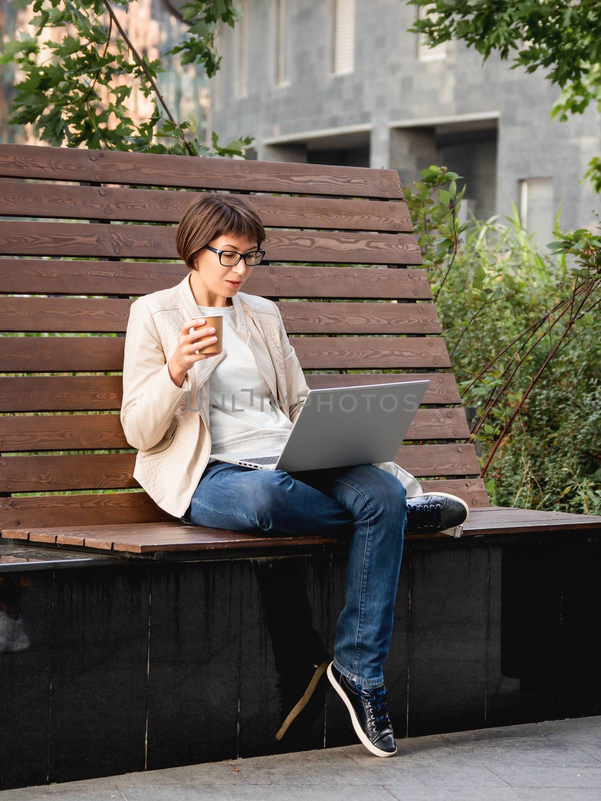 Freelance business woman sits in park with laptop and take away cardboard cup of coffee. Casual clothes, urban lifestyle of millennials. Working remotely.