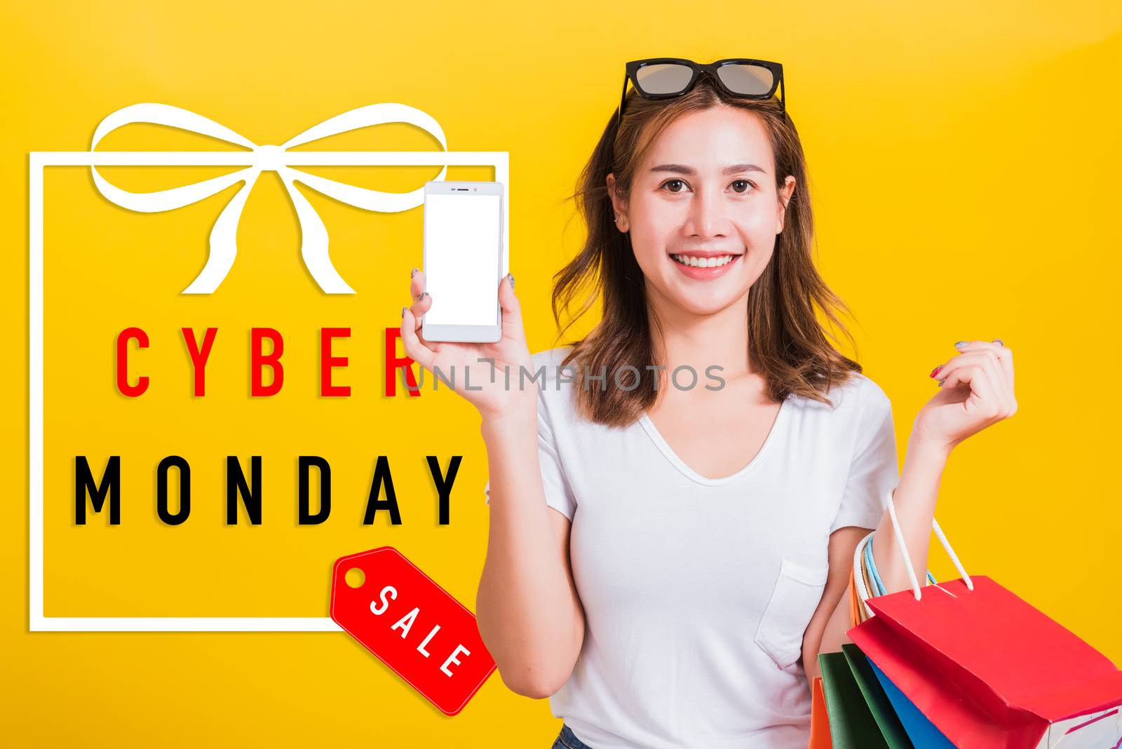 Portrait happy Asian beautiful young woman smile standing she holding shopping bags and show mobile phone blank screen with Cyber Monday text in gift box on side, studio shot on yellow background