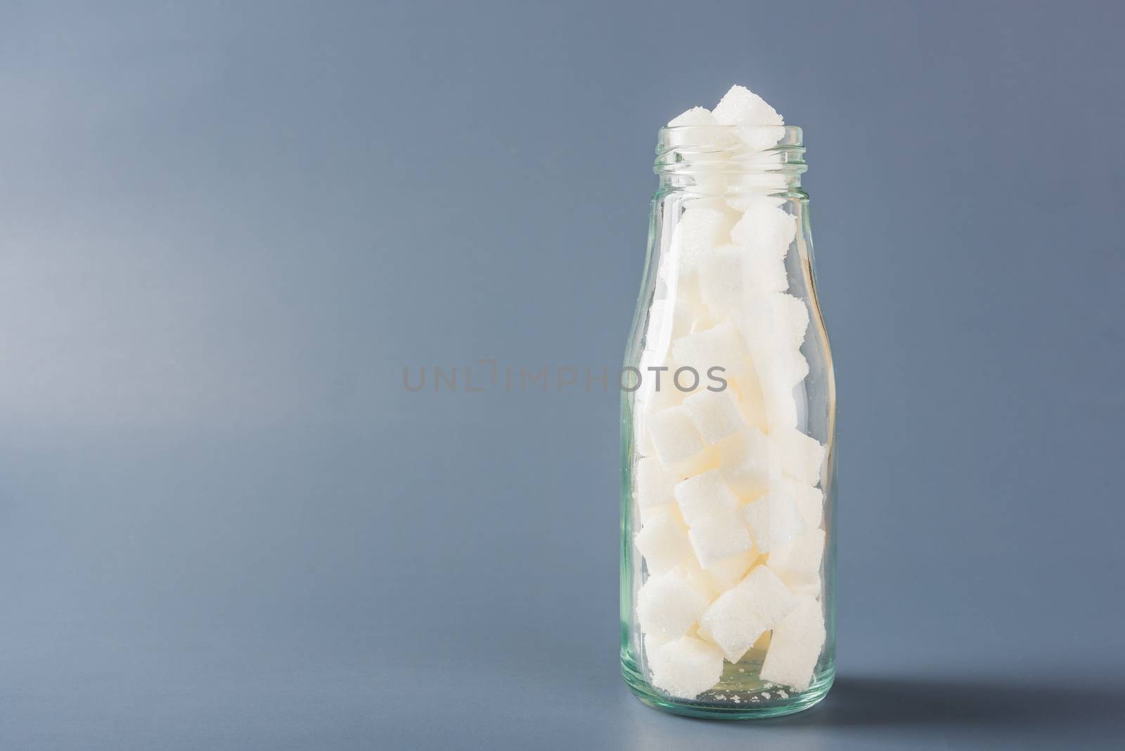 Glass bottle full of white sugar cube sweet food ingredient, studio shot isolated on gray background, health high blood risk of diabetes and calorie intake concept and unhealthy drink