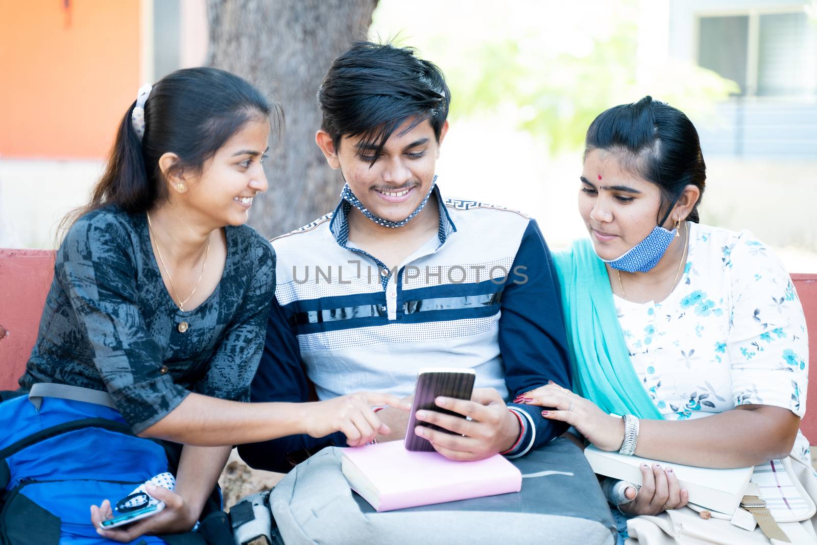 Three friends while sitting at college laughing by looking into mobile phone - concept of students internet, mobile addiction, online bullying or gossips at university campus by lakshmiprasad.maski@gmai.com