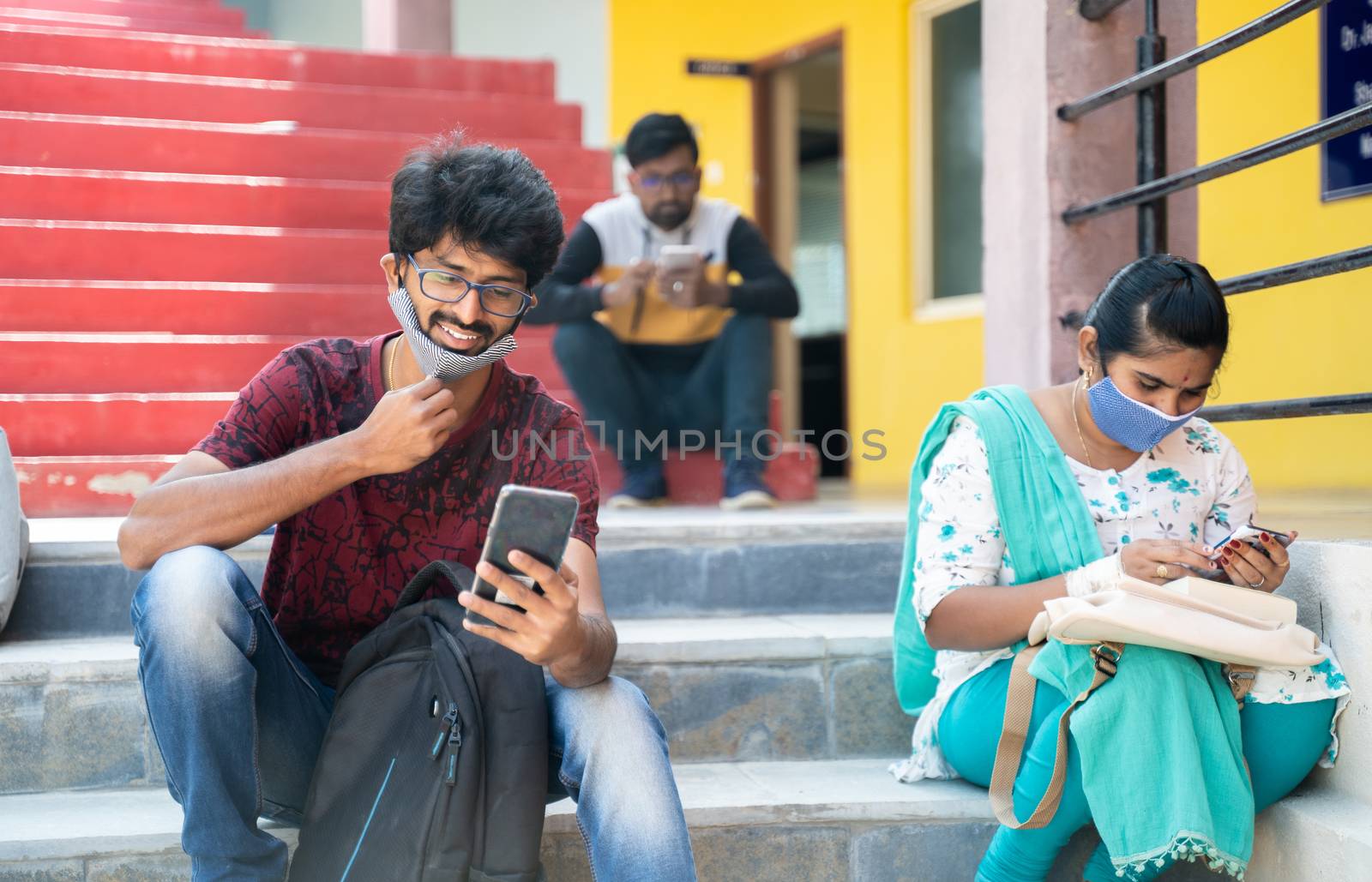 college students busy using mobile with face mask - happy millennial friends at university campus having fun during break - Concept of new normal lifestyle after college reopen. by lakshmiprasad.maski@gmai.com
