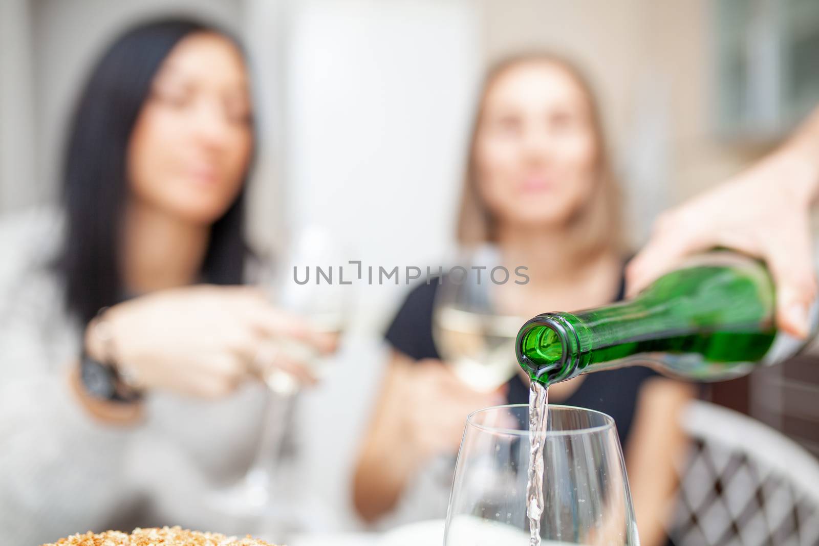 A male hand in the foreground pours wine into glasses. There are two female figures in the background. Friends meeting. Two unrecognizable girls drink wine in the home kitchen.