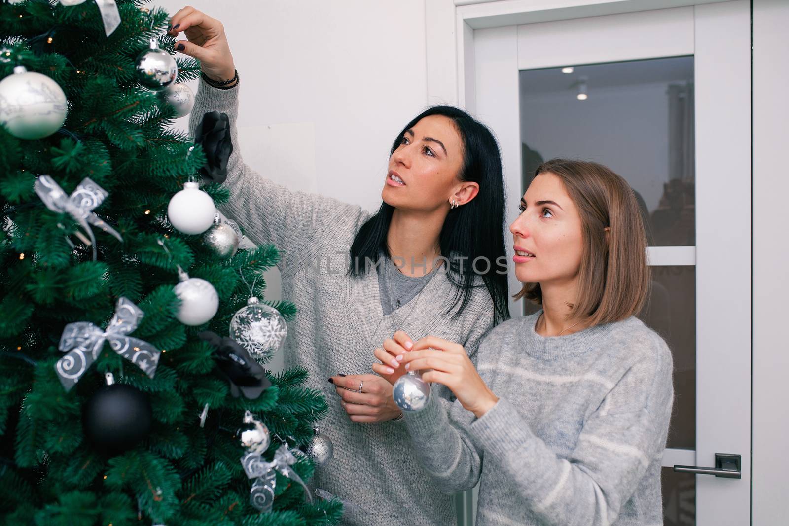 Two young women decorate the Christmas tree, preparing for the New Year's celebration. Friends decorate a Christmas tree. Beautiful girls are smiling and have fun during the Christmas holidays.