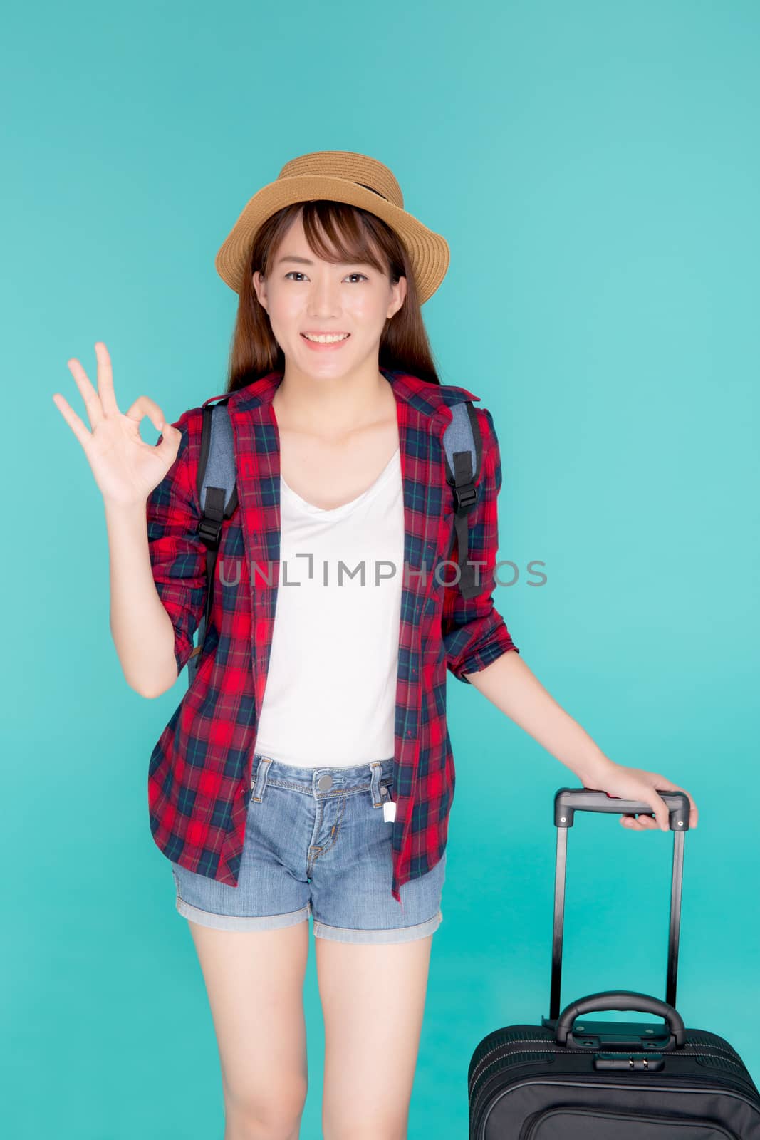 Beautiful young asian woman pulling suitcase isolated on blue background, asia girl cheerful holding luggage walking and gesture ok in vacation with excited, journey and travel concept.