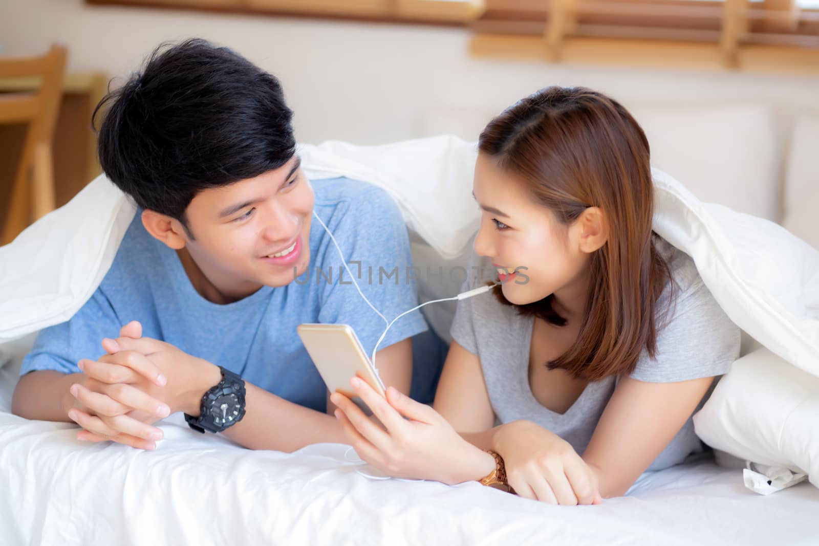 Young asian couple smile listen music with smart mobile phone on bed at bedroom with fun and enjoy, family sharing podcast or watching video from internet online with relax, lifestyle concept.