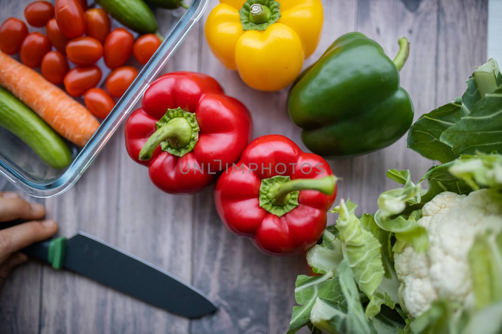 Colorful bell peppers, carrots, tomatoes, zucchini, and cauliflower on table from above. Female hand holding knife on wooden table.Vegan meal ingredients