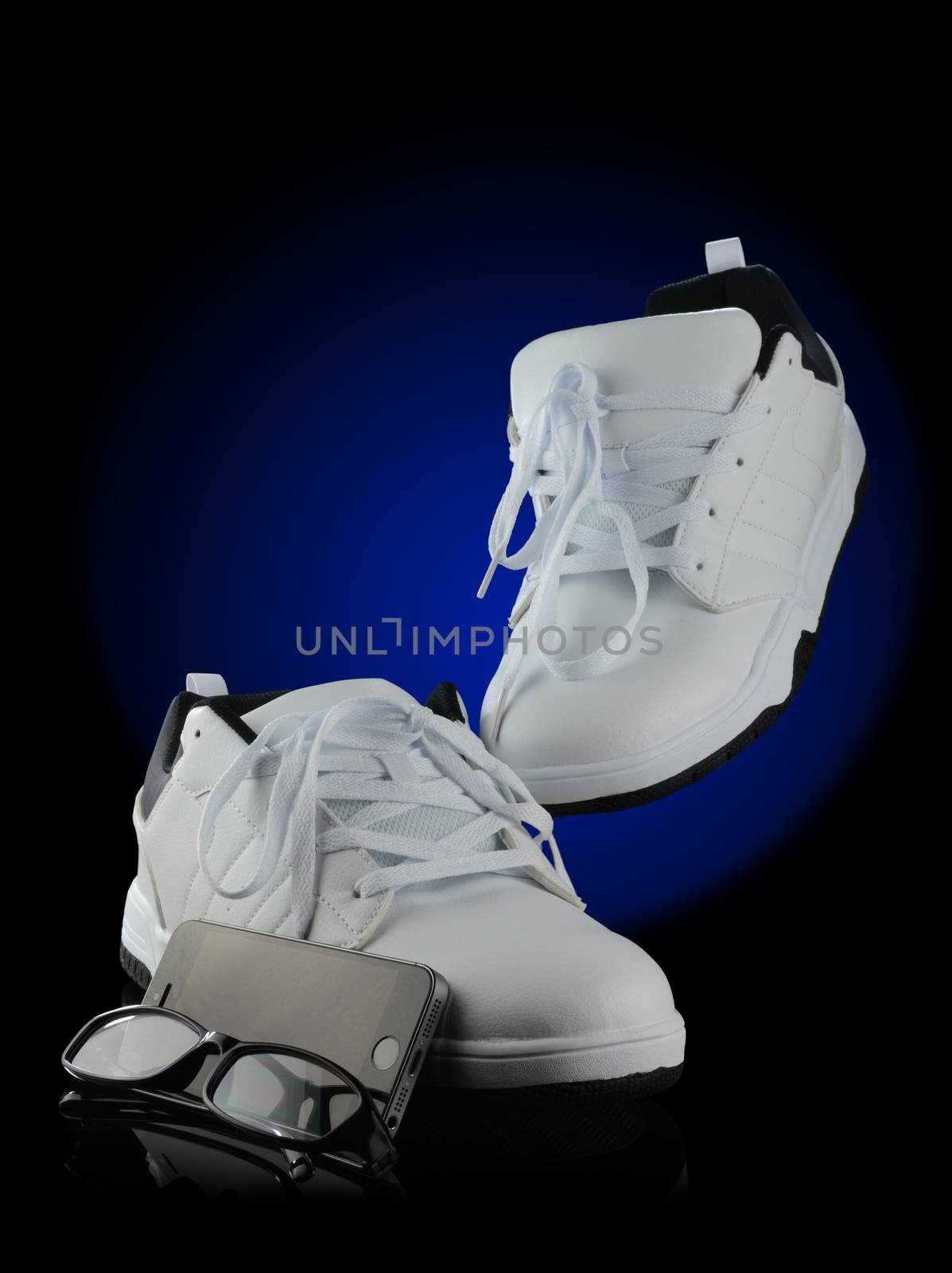 A poster style image of a pair of mens white street styled shoes with a cell phone and eye glasses over a black reflective background with a blue spot color for added depth.