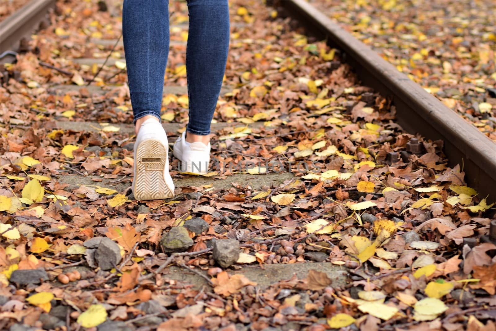 Walking legs wearing blue jeans and white shoes on autumn leaves and old railroad tracks