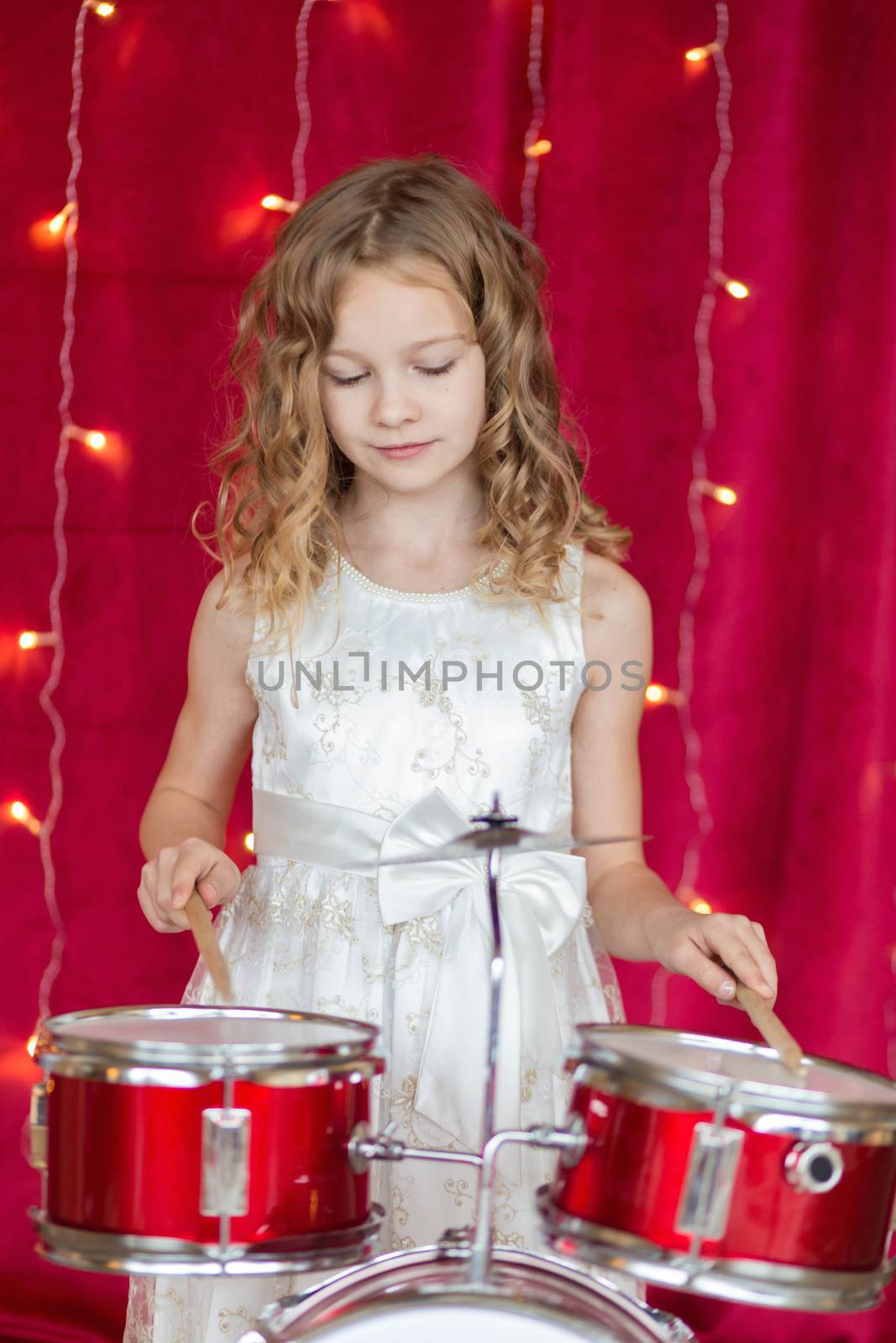 Little smiling girl plays on drums on red background with new year garlands.