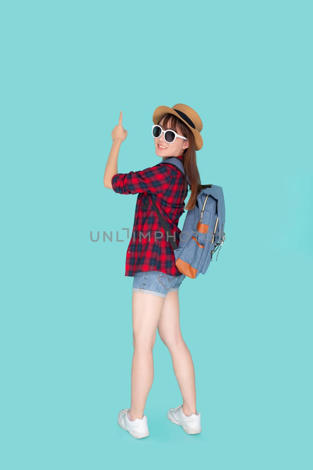 Back view beautiful portrait young asian woman wear hat and sunglasses smile excited enjoy travel summer trip in holiday isolated blue background, girl having backpack pointing something in vacation.
